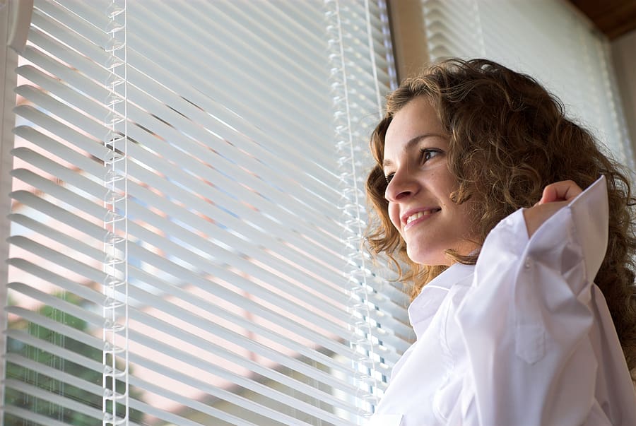 Smiling woman looking out of a window with blinds, illustrating the comfort and style brought by new window installations from Wagner. High-quality windows from brands like Wincore, Pella, and Velux provide energy efficiency, light, and a perfect fit for any space.